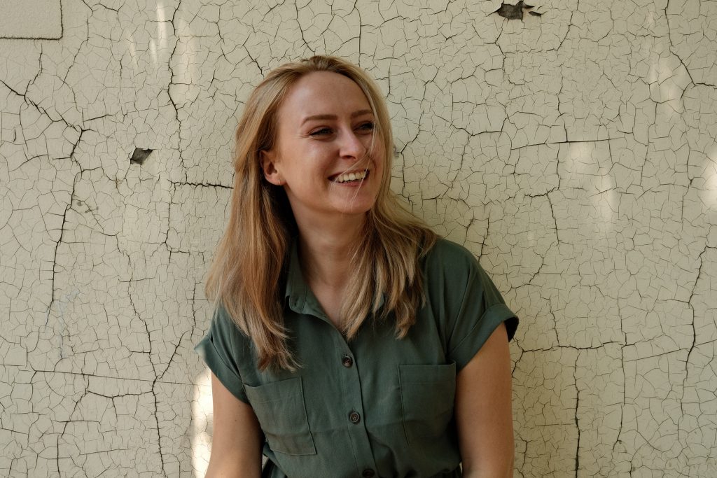 Portrait von Luise Wolff vor strukturierter Wand, lachend mit offenen Haaren und grünem Hemdkleid, Nahaufnahme mit natürlichem Licht