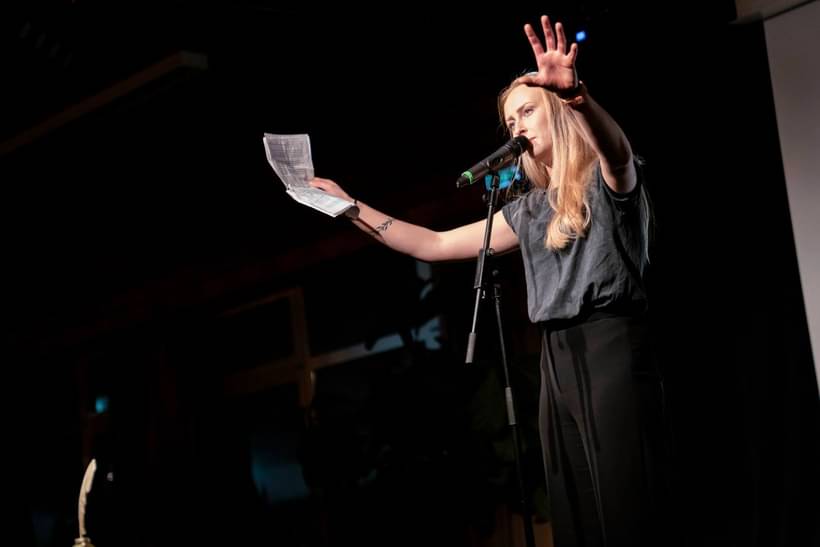Luise Wolff bei einem Poetry Slam Auftritt auf der Bühne, mit Mikrofon, erhobener Hand und Textblatt, in einem ausdrucksstarken Moment der Performance