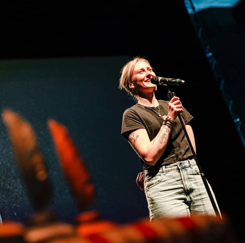 Luise Wolff lachend am Mikrofon bei einem Poetry Slam Auftritt, in Jeans und T-Shirt auf Bühne mit farbigen Requisiten im Vordergrund, 2025
