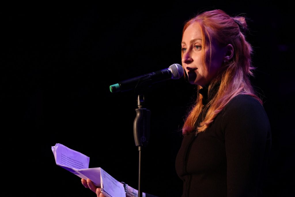 Luise Wolff bei einem Poetry Slam Auftritt mit Mikrofon und Textblatt in der Hand, im Scheinwerferlicht auf dunkler Bühne, 2024