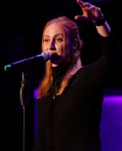 Luise Wolff bei einem Poetry Slam Auftritt auf der Bühne, mit Mikrofon und intensiver Gestik, Spotlight auf dunklem Hintergrund, 2024