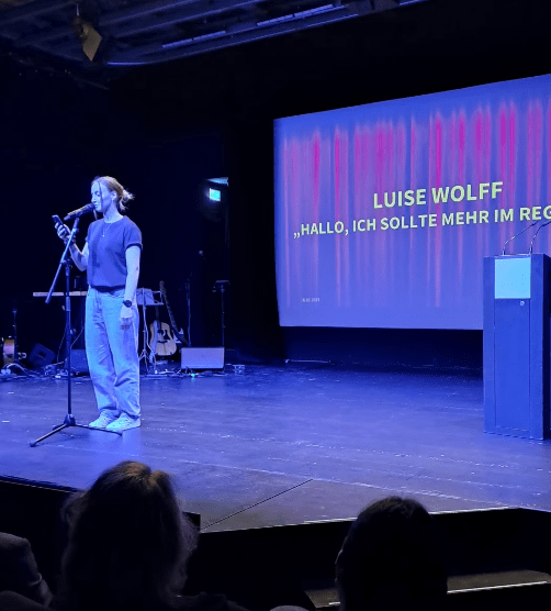 Luise Wolff steht auf einer Bühne am Mikrofon und liest einen Poetry Slam Text mit dem Titel "Hallo, ich sollte mehr im Regen stehen", der hinter ihr groß projiziert ist. Publikum sitzt im Vordergrund. Die Atmosphäre ist konzentriert und ruhig.